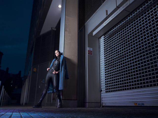 Image has a futuristic theme of dark blues and purples, featuring a model leaning against a pillar outside a covered carpark entrance