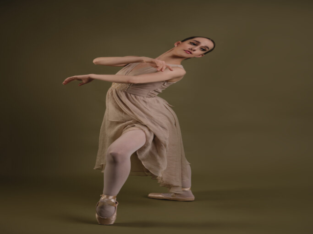 Image of Erica, a ballerina. A portrait of Erica in a beige dress in a dance pose, against a khaki background