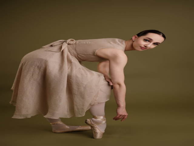 Image of Erica, a ballerina. A portrait of Erica in a beige dress in a dance pose, against a khaki background
