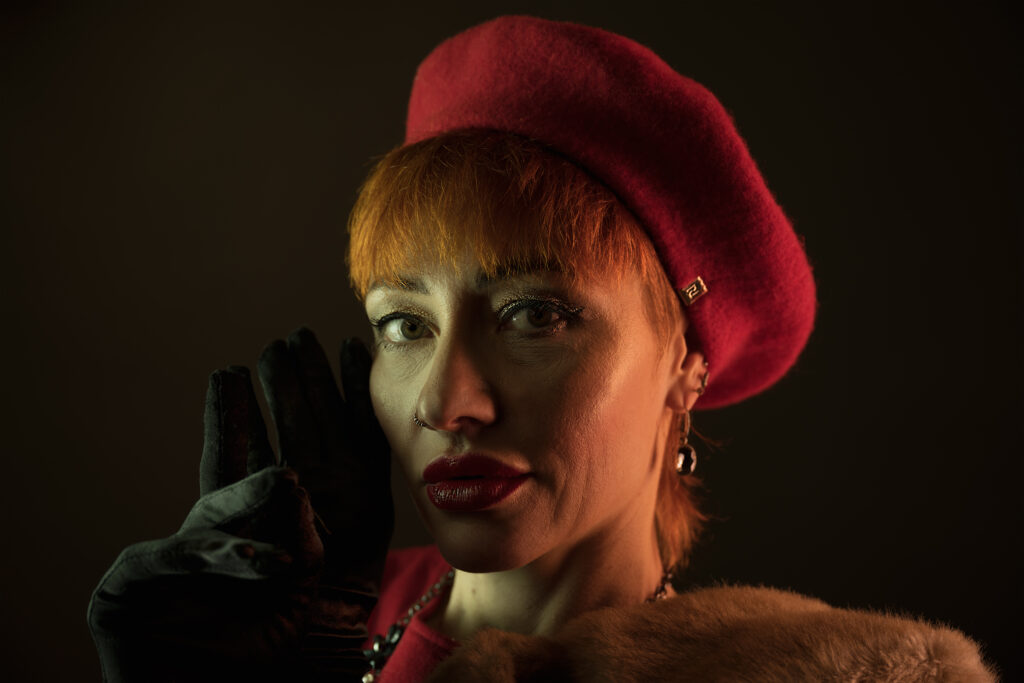 Image featuring a head and upper torso shot of Liliya, a model with red/orange hair in a faux fur coat, red dress, red beret and large jewellery.
