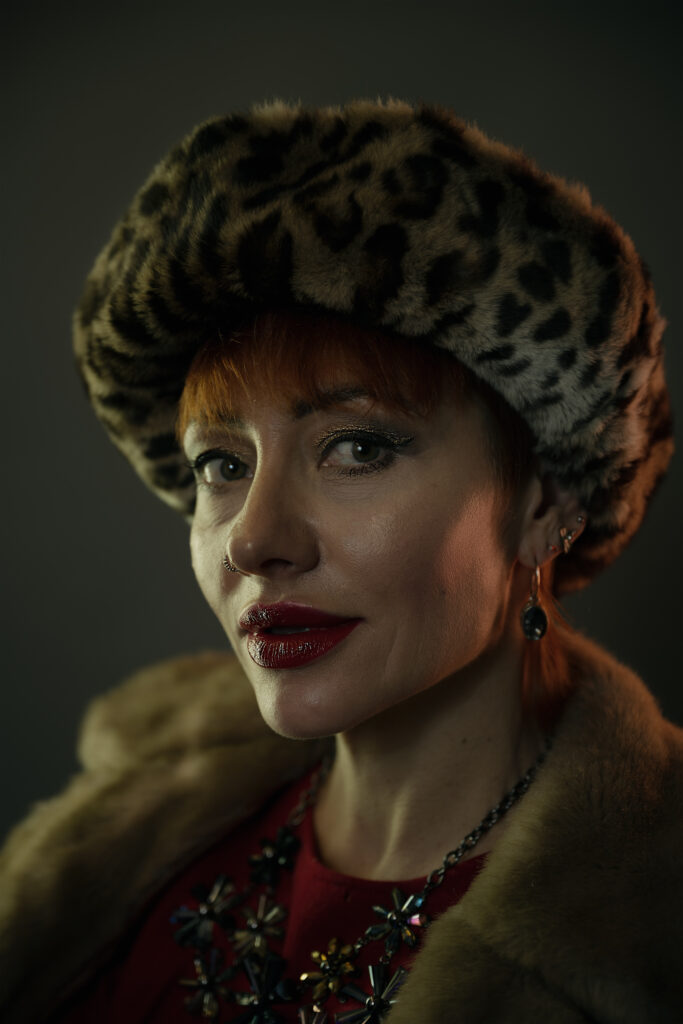 Image featuring a head and upper torso shot of Liliya, a model with red/orange hair in a faux fur coat, red dress, faux fur hat and large jewellery.