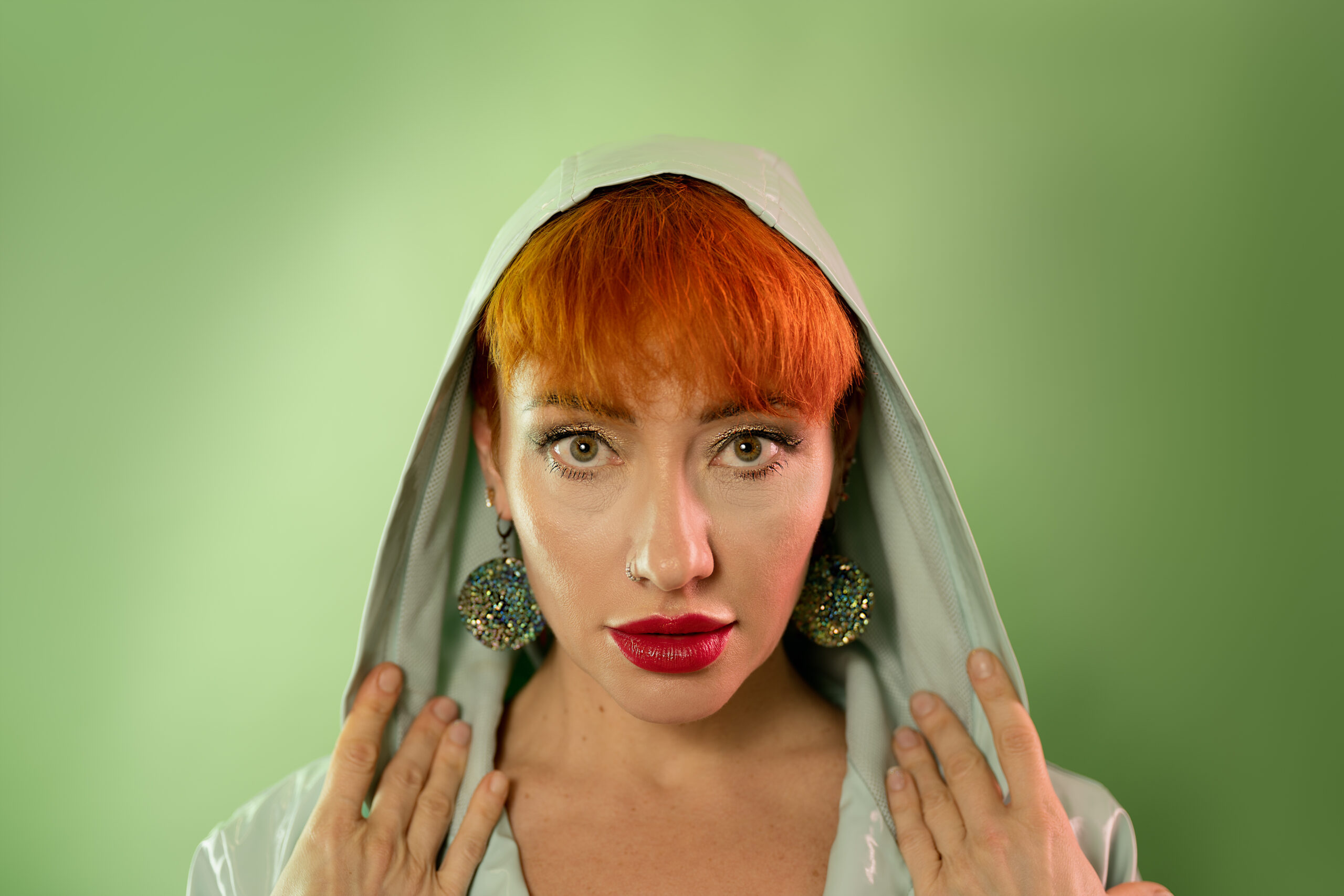 Image is green on green, featuring ahead and shoulders shot of Liliya, a model with red/orange hair in a green mac against a green background