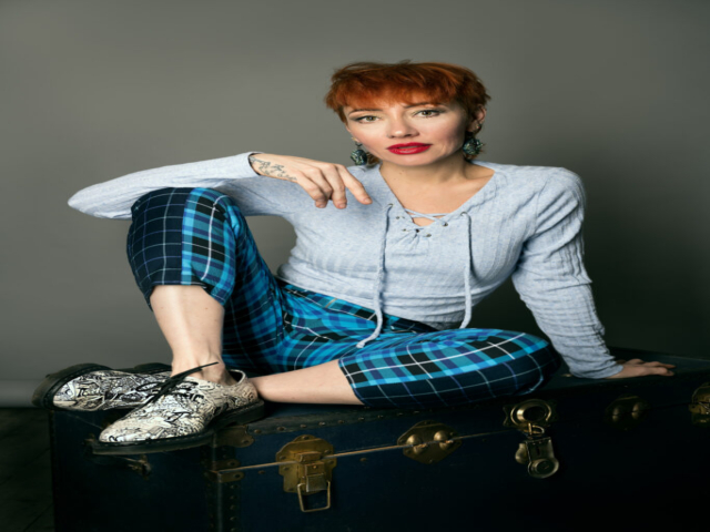 Image featuring Liliya, a model with red/orange hair wearing blue clothing with black and white shoes, sat on a blue chest, against a grey background,