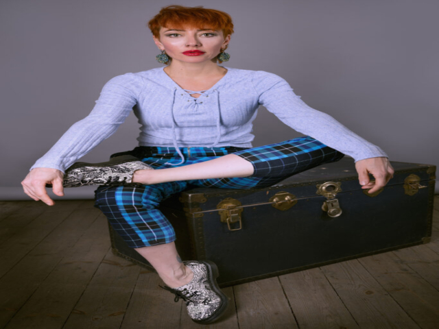 Image featuring Liliya, a model with red/orange hair wearing blue clothing with black and white shoes, sat on a blue chest, against a grey background,