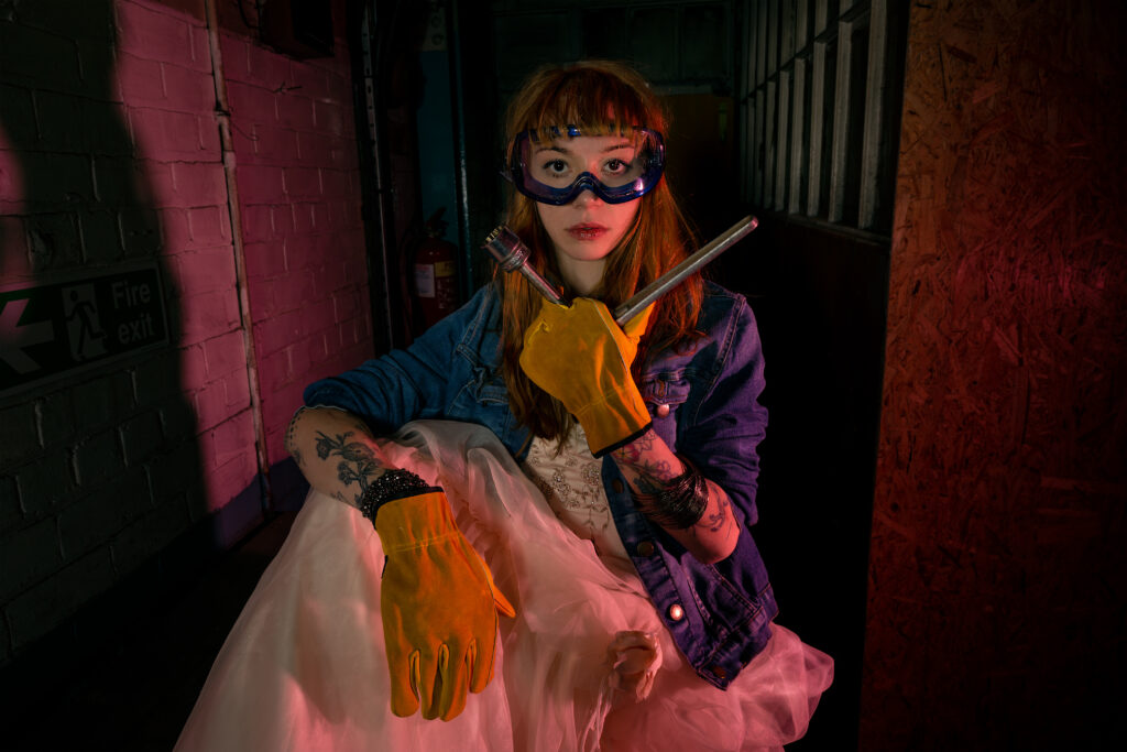 Featuring a caucasian female model, looking straight at camera. Wearing an ivory wedding dress, blue denim jacket, yellow leather gloves and blue safety goggles. Model is holding a wrench in a factory setting in red/purple light