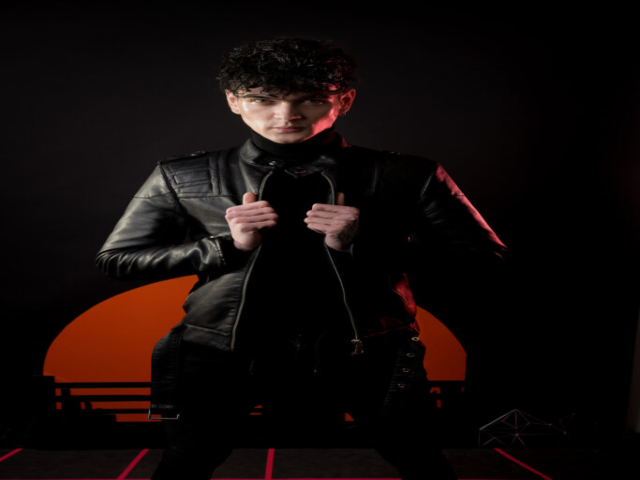 Male model in leather jacket, looking straight into the camera. He is stood in front of an orange striated sun and standing on a retrowave-inspired grid floor