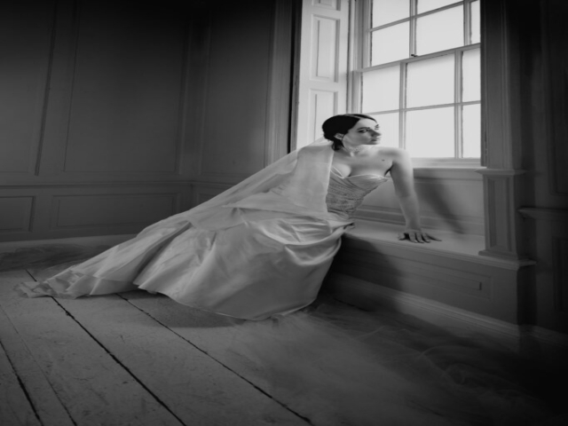 Black and white image of a caucasian female model sat in wedding gown, in a white studio against a large window