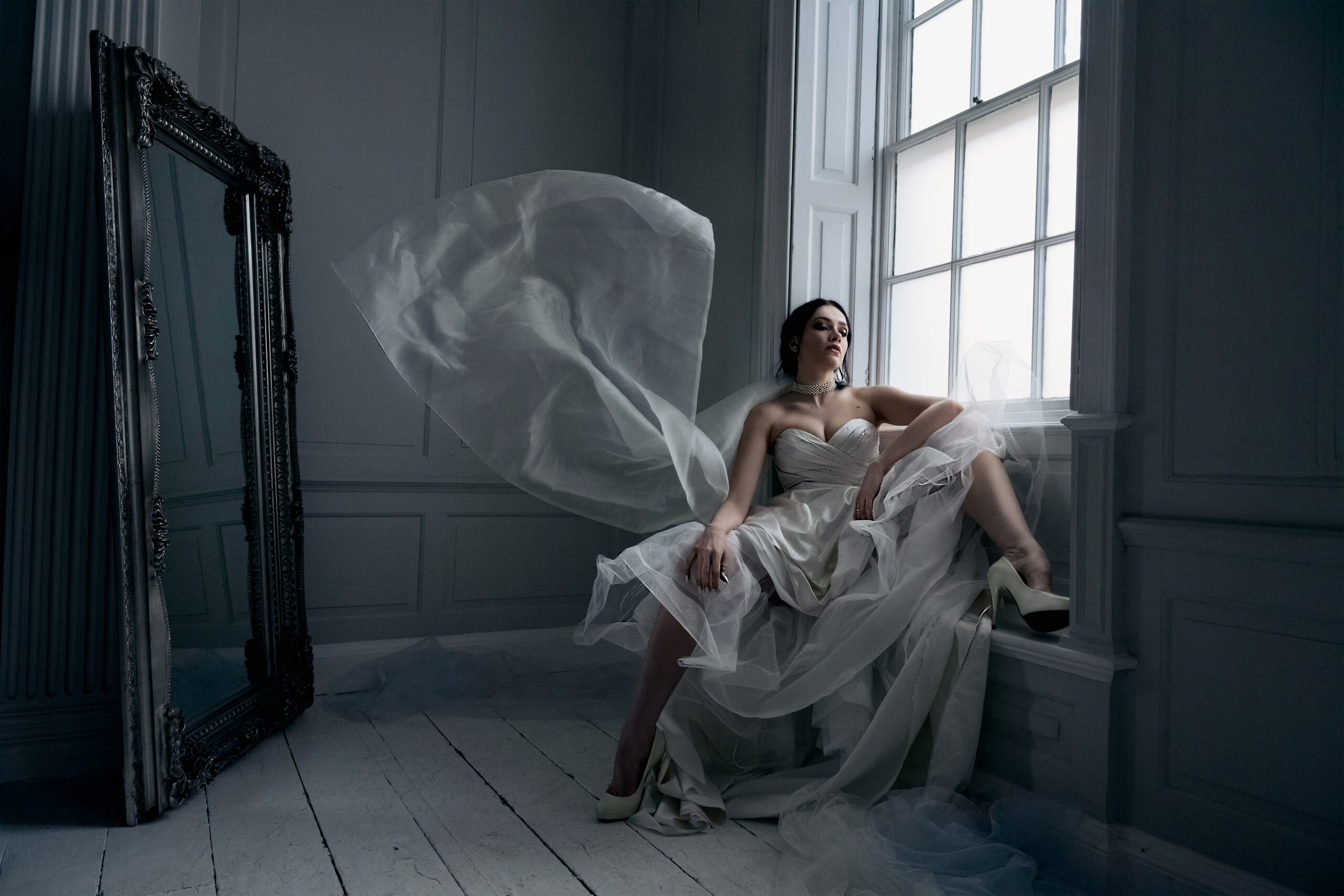 Female, caucasian model sat in wedding gown, with fabric floating mid-air, in a white studio against a large window