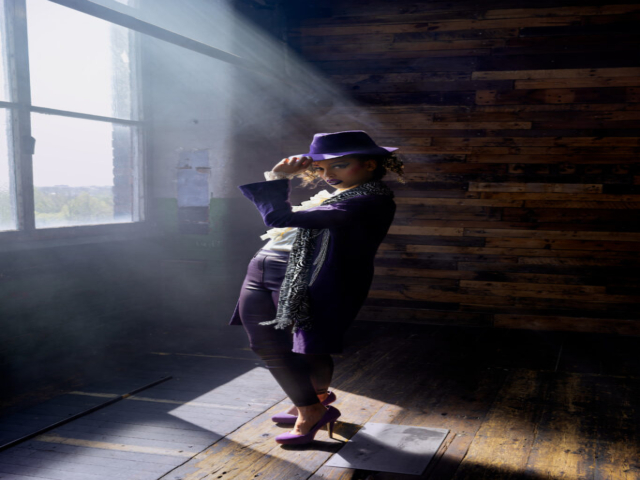 Image features a model in character as the musician Prince, standing in light shafts from the window to the left, looking at the camera while holding hat