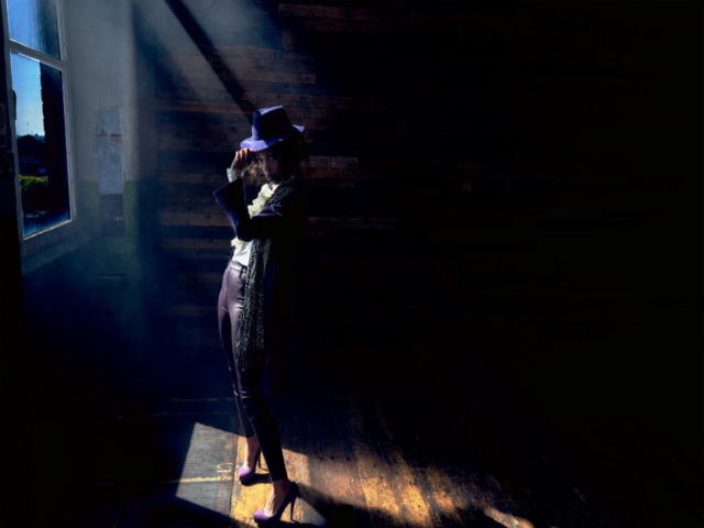 Image features a model in character as the musician Prince, looking at the camera. Model is in shafts of light from the windows to the left of the image