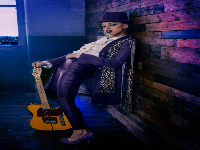 Image features a model in character as the musician Prince, stood against wall in a mill studio, with t-style guitar