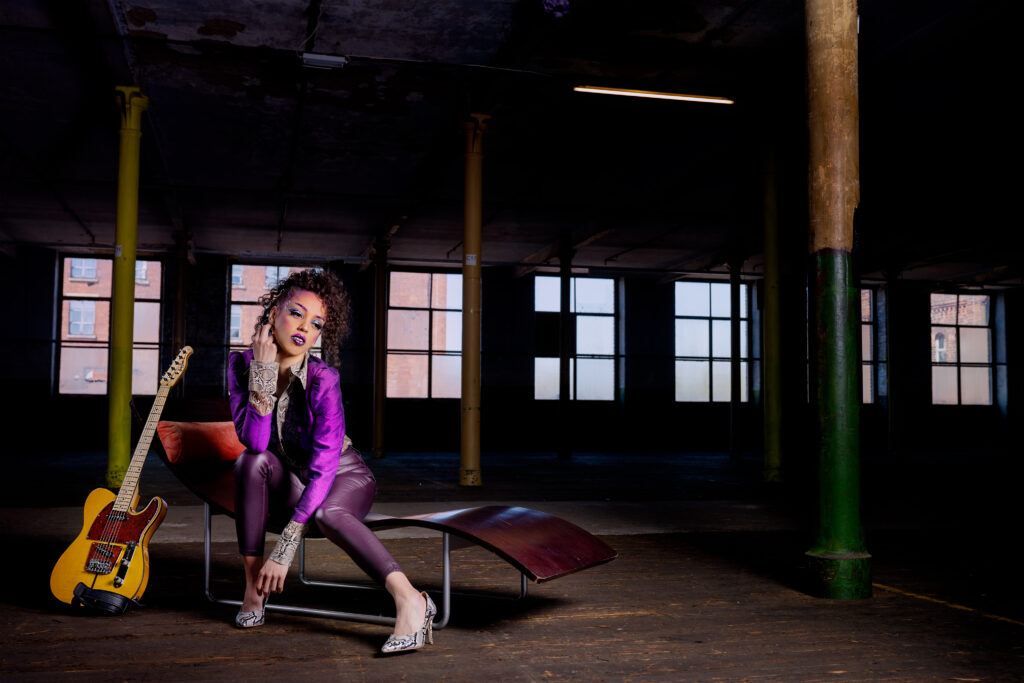 Image features a model in character as the musician Prince, say on a 1970s wooden chaise long next to replica t-style guitar in a mill studio