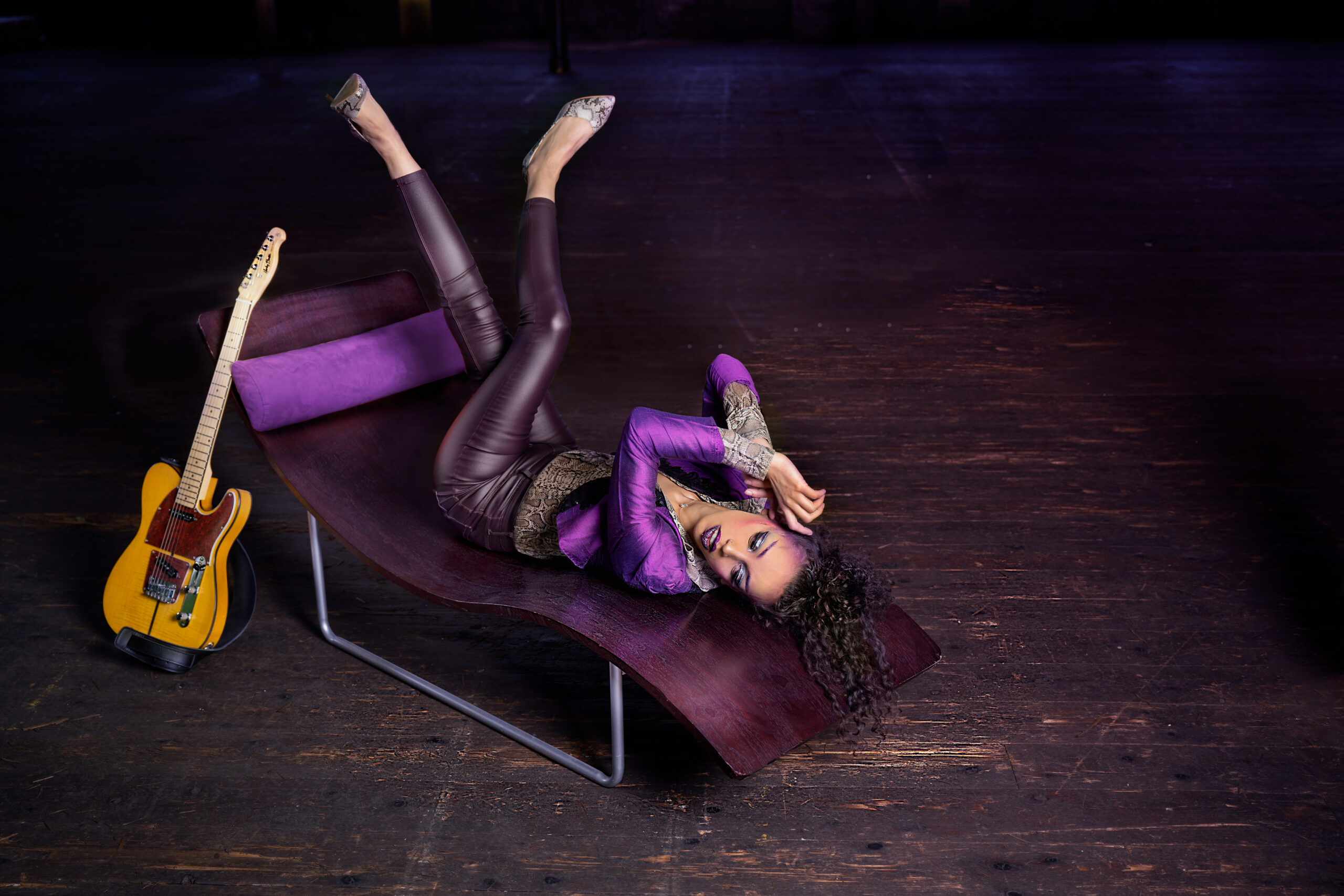Image features a model in character as the musician Prince, lying on a 1970s wooden chaise long next to replica t-style guitar in a mill studio