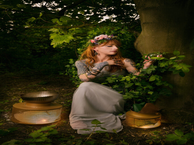 Tattooed model seated in a forest is dressed with flower headpiece, holding a crystal ball in one hand. She's entwined in leaves and two lamps sit by her feet.