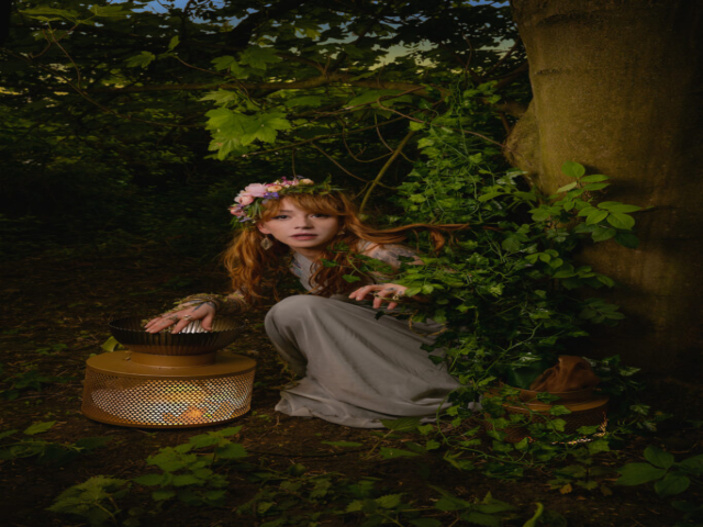 Model is dressed as a fairy princess with floral headpiece. She is crouching as if caught surprised mid action. Her right hand is hovering over a golden bowl over a lit lantern. She is next to a tree and partly covered in leaves within the woodland
