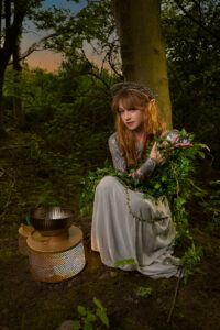 Model is dressed as a fairy princess with silver headpiece in woodland. She is crouching, holding two crystal balls in her right hand to her left shoulder