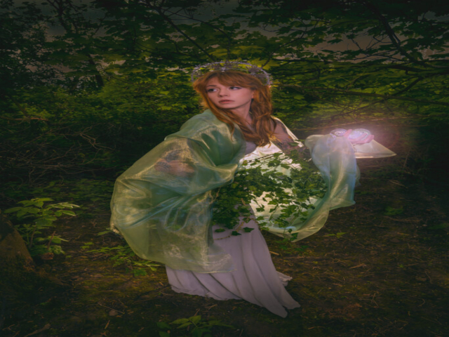Model in a forest is dressed with silver headpiece and elf ears, holding a crystal ball in one hand. She's entwined in leaves and vine and covered in a shawl. Light emanates from two balls she's holding in her hand.