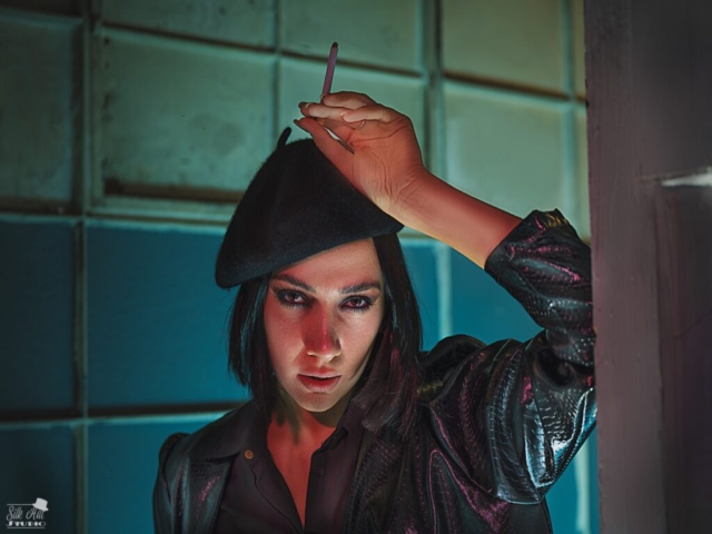 Features a female model leaning against a wall in a corridor. The model is wearing black with a shiny coat, black beret and holding a (fake) cigarette to the beret. The background walls have been lit in blue and green.