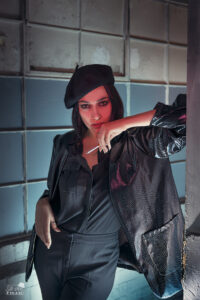 Features a female model leaning against a wall in a corridors. The model is wearing black with a shiny coat, black beret and holding a (fake) cigarette to the beret. The background walls have been lit in blue.