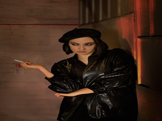 Features a female model leaning against a wall in a corridors. The model is wearing black with a shiny coat, black beret and holding a (fake) cigarette to her side.
