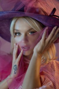 Headshot of Katey, blonde, female model with pink makeup on a pink background. Katey is wearing a purple hat and holding a pink gauze up to the lower half of her face