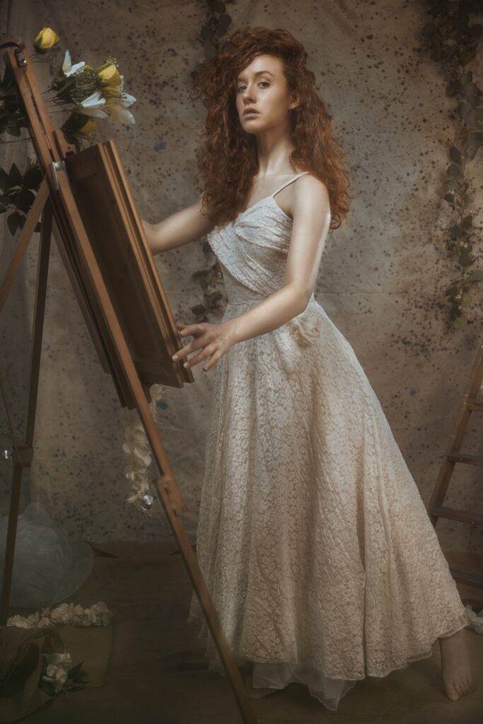Image is a fine art image, featuring a Caucasian woman with red hair wearing a vintage lace wedding dress. She is standing posed next to an easel with a framed image and flowers on it