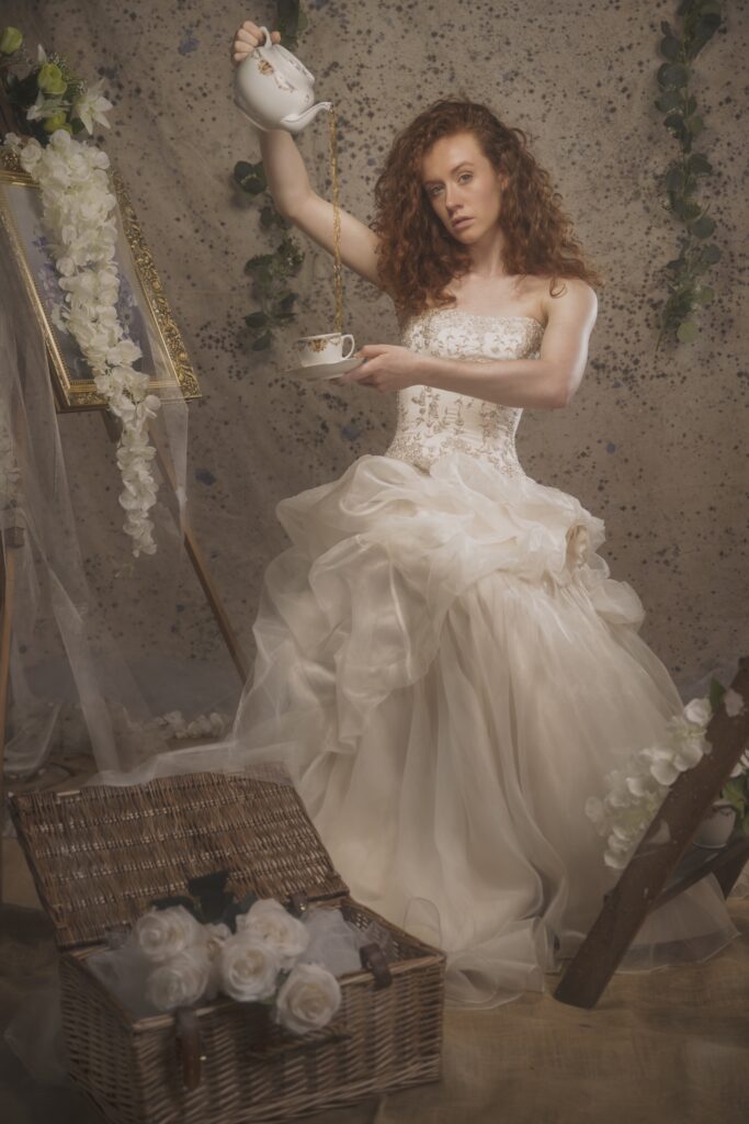 Image is a fine art image, featuring a Caucasian woman with red hair wearing a vintage wedding dress. She is pouring tea from a teapot at height into a cup