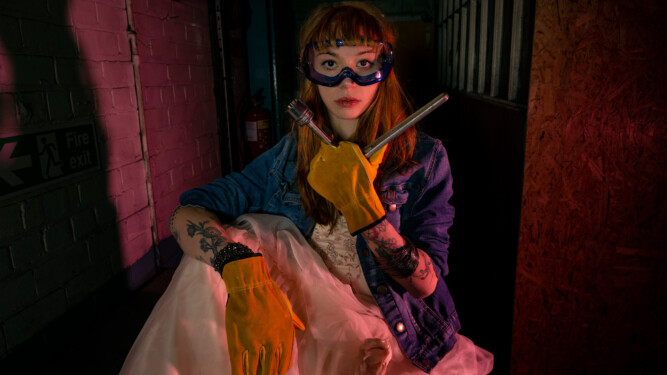 Featuring a caucasian female model, looking straight at camera. Wearing an ivory wedding dress, blue denim jacket, yellow leather gloves and blue safety goggles. Model is holding a wrench in a factory setting in red/purple light