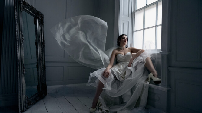 Female, caucasian model sat in wedding gown, with fabric floating mid-air, in a white studio against a large window