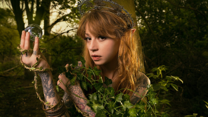 Tattooed model in a forest is dressed with silver headpiece and elf ears, holding a crystal ball in one hand. She's entwined in leaves and vine.